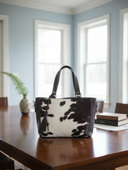 Black and white cowhide handbag on a wooden table with a light blue wall and windows in the background.
