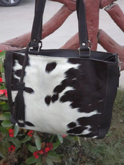 Black and white cowhide patterned handbag with leather straps against a blurred outdoor background.