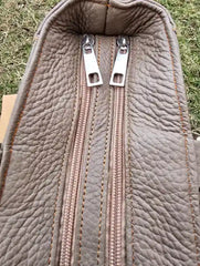 Close-up of a textured brown leather bag with two zippers on a grassy background.